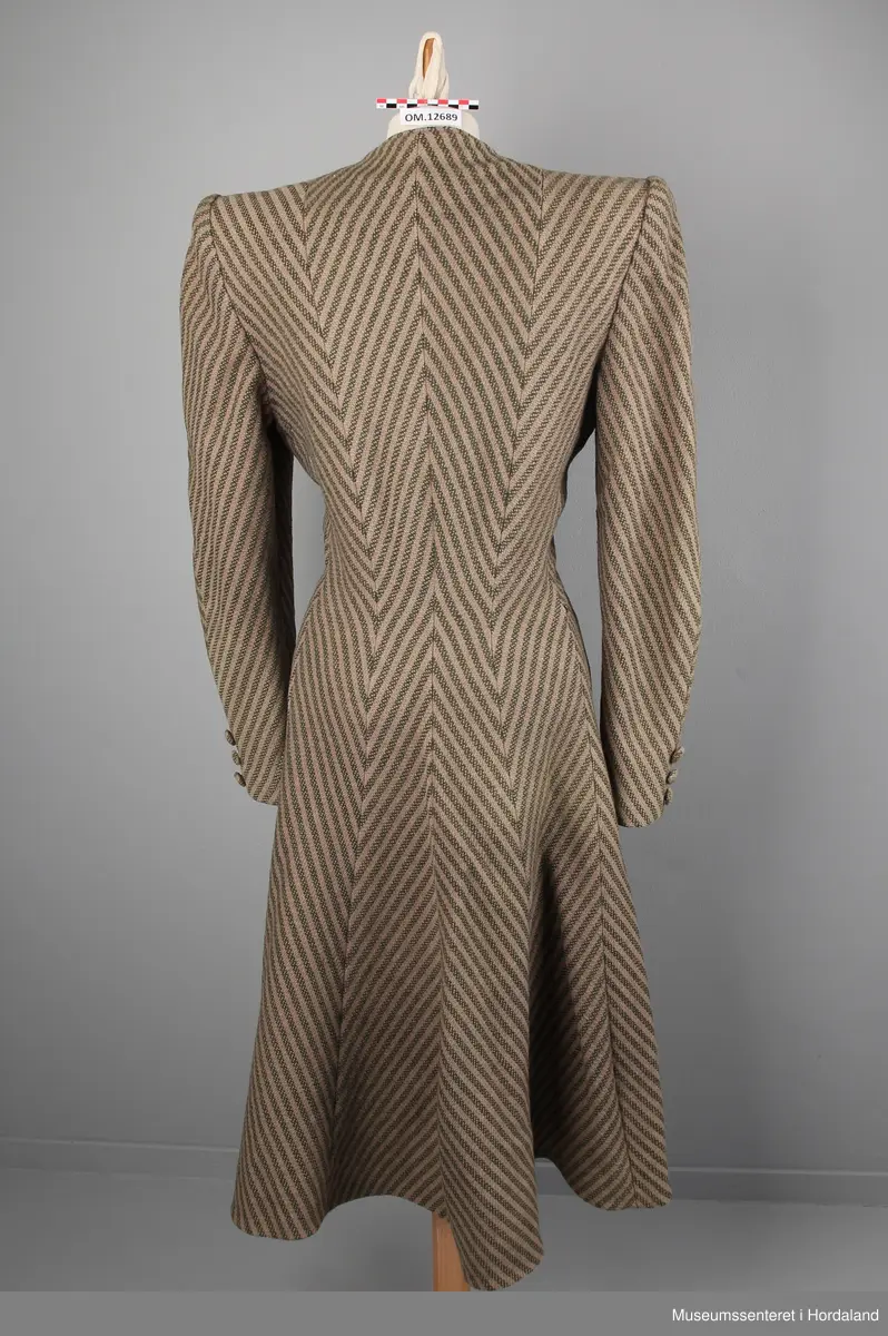 Kåpa er sydd saman av 10 bredder (skråklipte), der alle stripene møtes rett i alle saumane. Fasongsydd og sterkt innsvinga i livet. Slag, men ikkje krage. Trekte knappar - 9 stk i same stoff som kåpa. Knapphola ligg i ein saum - altså ikkje klipte opp og sydde. To stikklommer på kvar side framme i saumen. Tre trekte knappar framme på ermene. Fora med kunstsilkestoff. På innsida nede har kåpa skoning (forsterking). Store skulderputer.