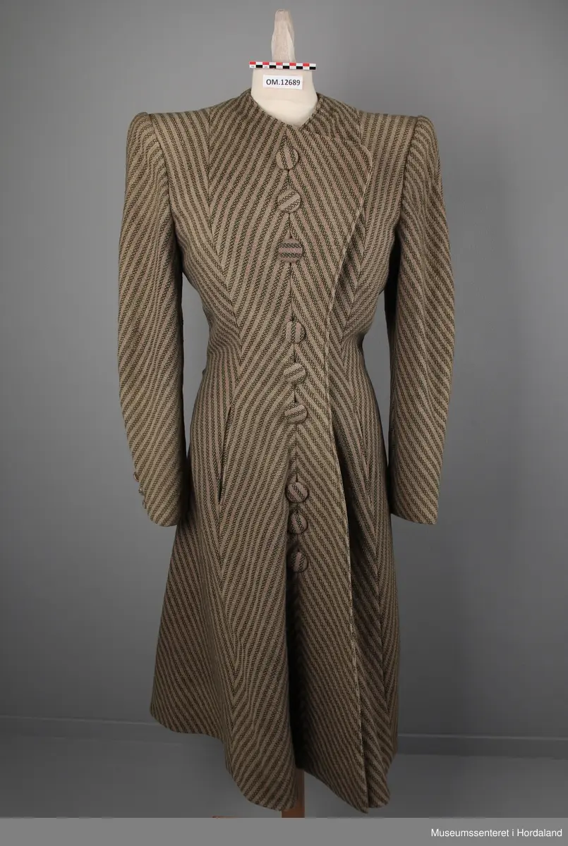 Kåpa er sydd saman av 10 bredder (skråklipte), der alle stripene møtes rett i alle saumane. Fasongsydd og sterkt innsvinga i livet. Slag, men ikkje krage. Trekte knappar - 9 stk i same stoff som kåpa. Knapphola ligg i ein saum - altså ikkje klipte opp og sydde. To stikklommer på kvar side framme i saumen. Tre trekte knappar framme på ermene. Fora med kunstsilkestoff. På innsida nede har kåpa skoning (forsterking). Store skulderputer.