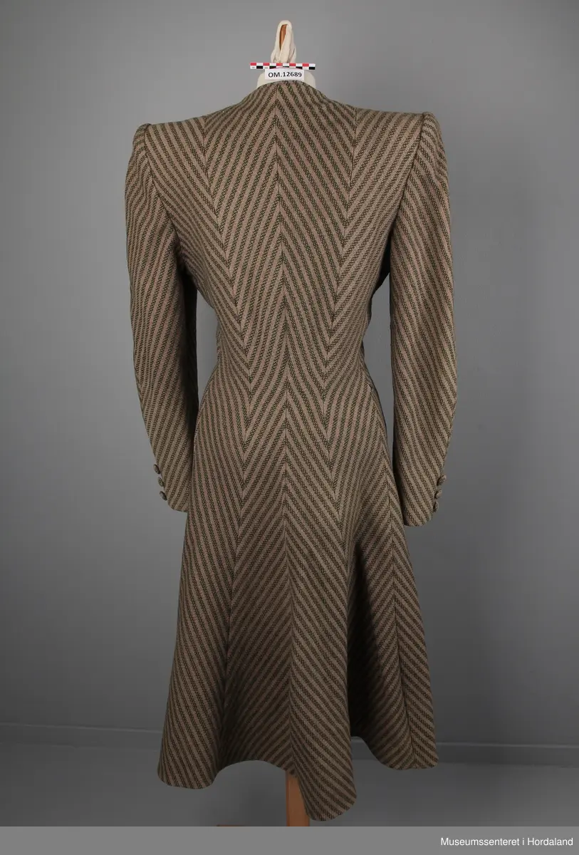 Kåpa er sydd saman av 10 bredder (skråklipte), der alle stripene møtes rett i alle saumane. Fasongsydd og sterkt innsvinga i livet. Slag, men ikkje krage. Trekte knappar - 9 stk i same stoff som kåpa. Knapphola ligg i ein saum - altså ikkje klipte opp og sydde. To stikklommer på kvar side framme i saumen. Tre trekte knappar framme på ermene. Fora med kunstsilkestoff. På innsida nede har kåpa skoning (forsterking). Store skulderputer.