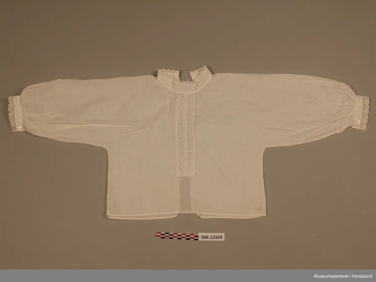 Barnebunadsskjorte i kvitt bomullsstoff. Lange armar. Heilt open bak med to knappar og knapphol oppe. Ingen festing i nedre halvdel. Linning i halsen. Til stolpemønster framme er det sydd fast ein blonde. Det er ein smal blonde på krage og armar. Skjorta er rynka ved armmansjettane, elles ingen rynking. Enkelt snitt utan kiler.