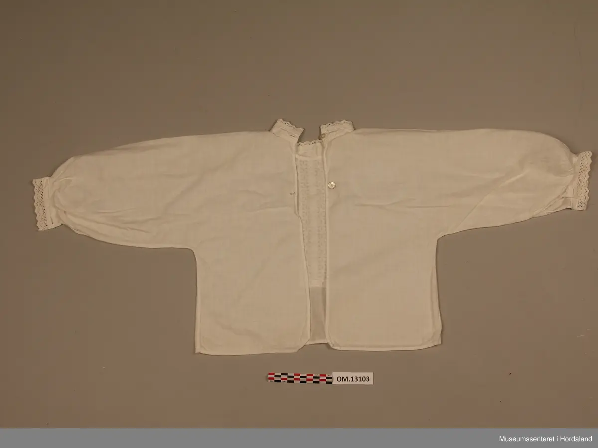 Barnebunadsskjorte i kvitt bomullsstoff. Lange armar. Heilt open bak med to knappar og knapphol oppe. Ingen festing i nedre halvdel. Linning i halsen. Til stolpemønster framme er det sydd fast ein blonde. Det er ein smal blonde på krage og armar. Skjorta er rynka ved armmansjettane, elles ingen rynking. Enkelt snitt utan kiler.