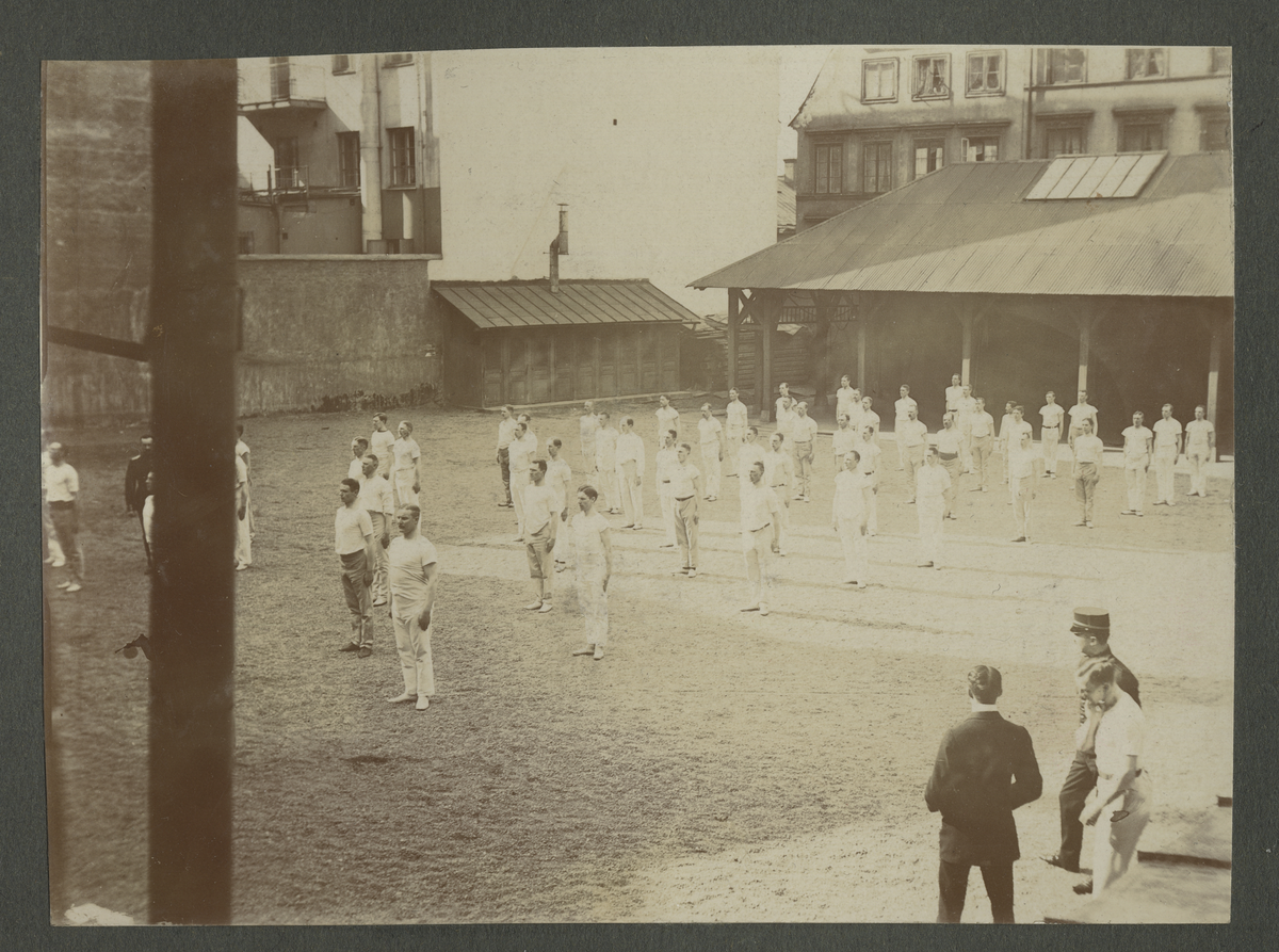 En samling män iklädda träningskläder springer på led om två runt en grusplan vid Gymnastiska central institutet (GCJ 1911-1912. I förgrunden syns två män varav en ler glatt mot kameran.