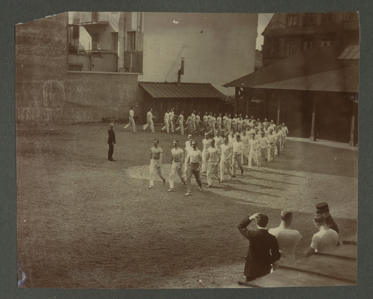 Gymnastiska central institutet. Män utövar gymnastik på gårdplan omgiven av hus under 1911-1912, alla klädda i vita träningskläder. Bredvid står tre män och inspekterar.