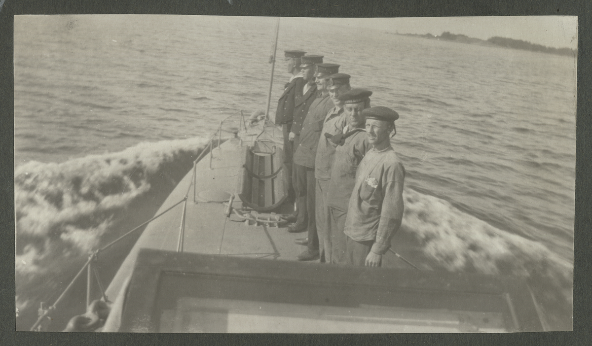 Karlskrona 1914 - Man står på ubåt och signalera med flaggor i händerna. Tre andra matroser står i bakrunden på ubåtsdäck.