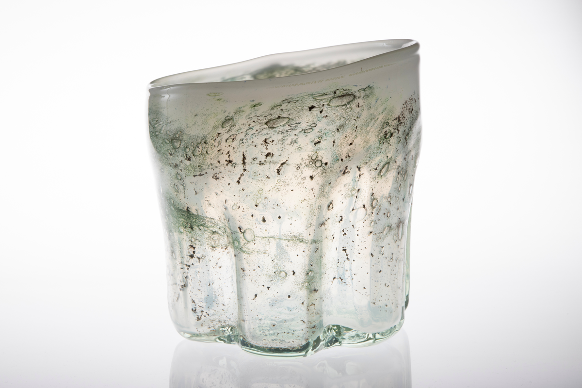 Irregulær sylinderformet vase i halvgjennomskinnelig melkehvitt glass. Ujevn munningsrand. Korpus er derkorert med metallspon, luftblærer og blå-, sort- og grønnfargede partier.