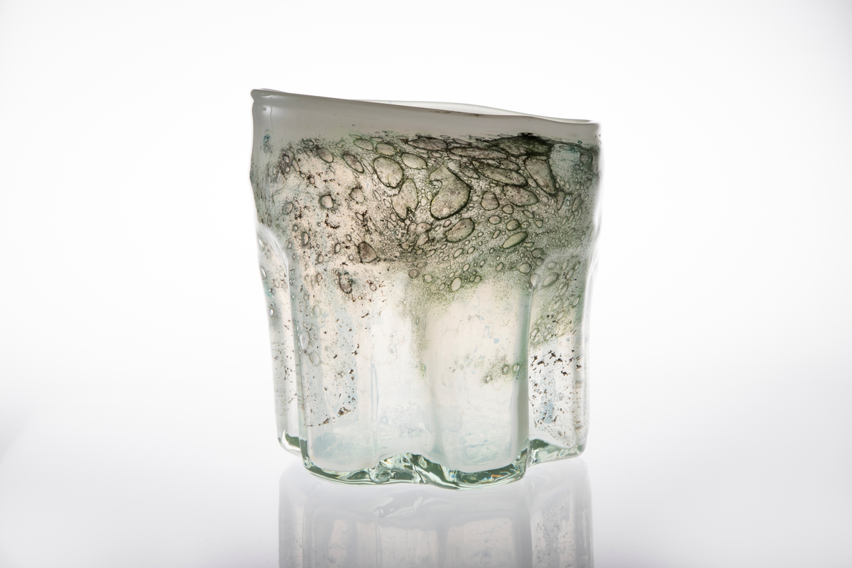 Irregulær sylinderformet vase i halvgjennomskinnelig melkehvitt glass. Ujevn munningsrand. Korpus er derkorert med metallspon, luftblærer og blå-, sort- og grønnfargede partier.