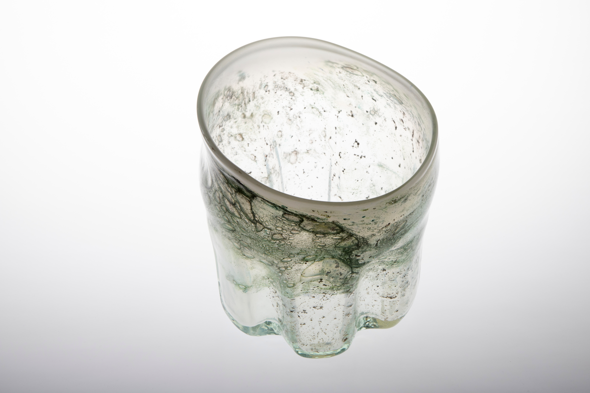 Irregulær sylinderformet vase i halvgjennomskinnelig melkehvitt glass. Ujevn munningsrand. Korpus er derkorert med metallspon, luftblærer og blå-, sort- og grønnfargede partier.