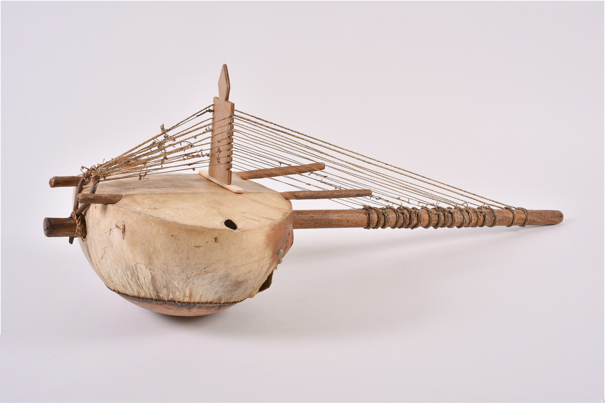 Harpe bestående av en kalebass trukket med skinn. Strenger festet i en lang pinne i tre, trukket i spenn over en bru over kalebassen og festet i ring av metall i enden. Skinnet festet med nagler av metall.