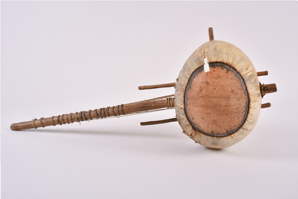 Harpe bestående av en kalebass trukket med skinn. Strenger festet i en lang pinne i tre, trukket i spenn over en bru over kalebassen og festet i ring av metall i enden. Skinnet festet med nagler av metall.