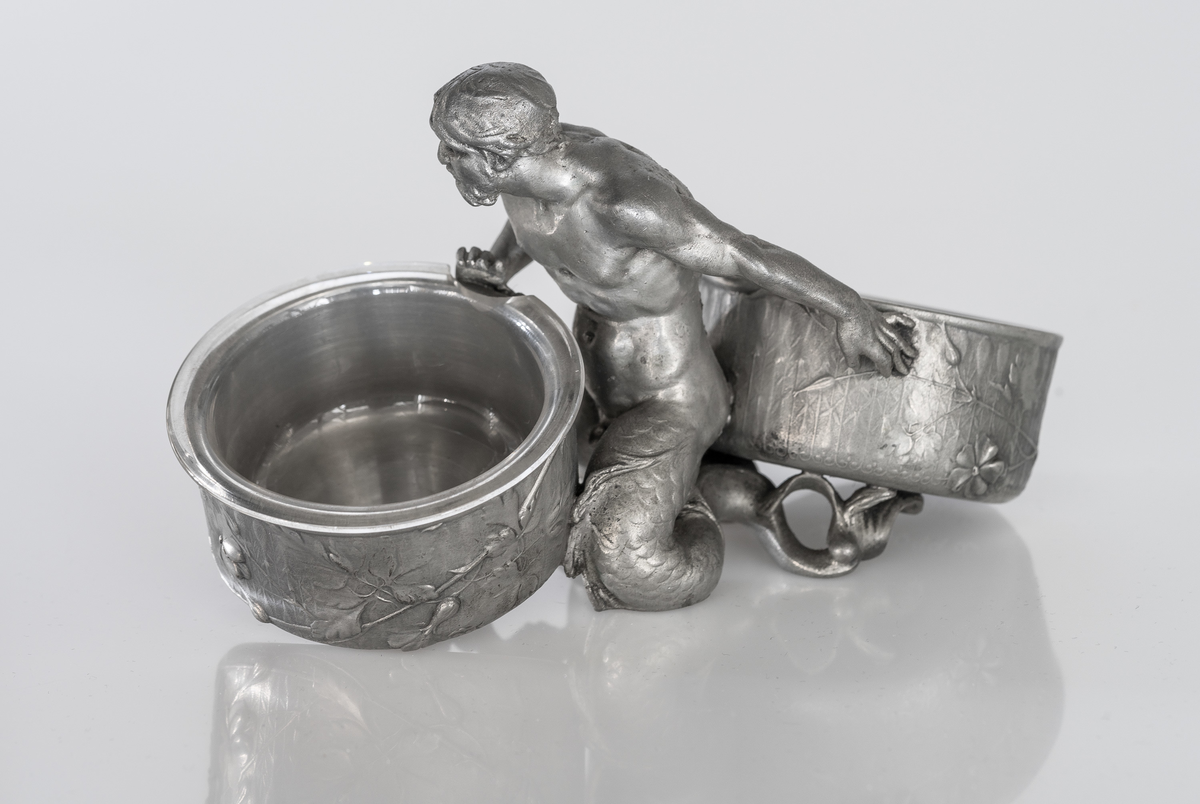 Saltkar i tinn, utformet som knestående havmann med bar overkropp og todelt fiskehale Hver av finnene støtter opp hver sitt kar, henholdsvis for salt og pepper, dekorert med storkenebbsplanter i svakt relieff.