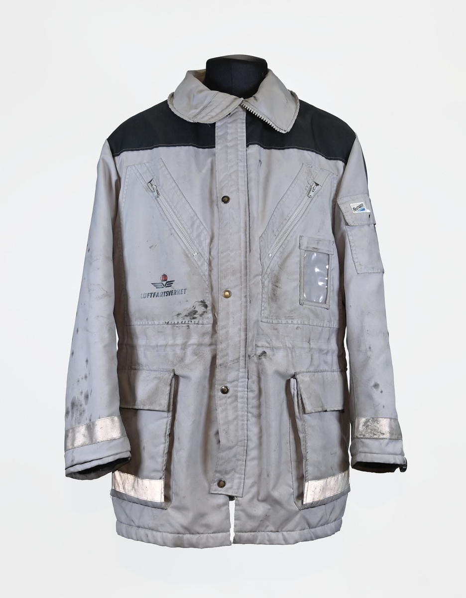 Grå varseljacka med svart foder tillhörande anställd på Luftfartsverket. Jackan har flera fickor som stängs med dragkedja eller kardborreband, samt en innerficka på vänster sida. Jackan är försedd med reflexer på båda ärmslut och båda framfickor. Luftfartsverkets logga sitte på höger bröst och ryggen. I jackan sitter en lapp med tvättanviselser. Jackan stängs med dragkedja i plast och sex stycken tryckknappar. Det finns även tryckknappar runt kragen för en eventuell luva som saknas. Jackan är väl använd och har flertalet smuts- och oljefläckar.

Jackan är i storlek L (54-56).