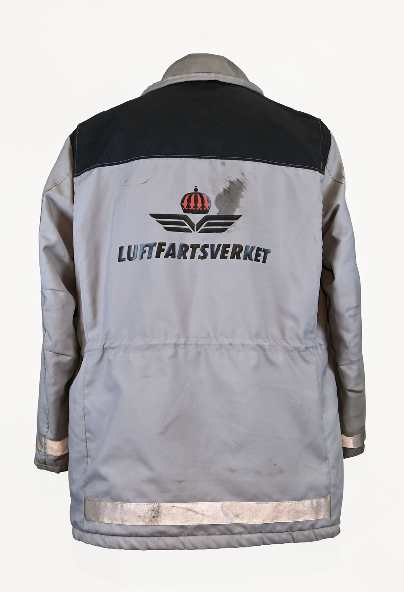 Grå varseljacka med svart foder tillhörande anställd på Luftfartsverket. Jackan har flera fickor som stängs med dragkedja eller kardborreband, samt en innerficka på vänster sida. Jackan är försedd med reflexer på båda ärmslut och båda framfickor. Luftfartsverkets logga sitte på höger bröst och ryggen. I jackan sitter en lapp med tvättanviselser. Jackan stängs med dragkedja i plast och sex stycken tryckknappar. Det finns även tryckknappar runt kragen för en eventuell luva som saknas. Jackan är väl använd och har flertalet smuts- och oljefläckar.

Jackan är i storlek L (54-56).