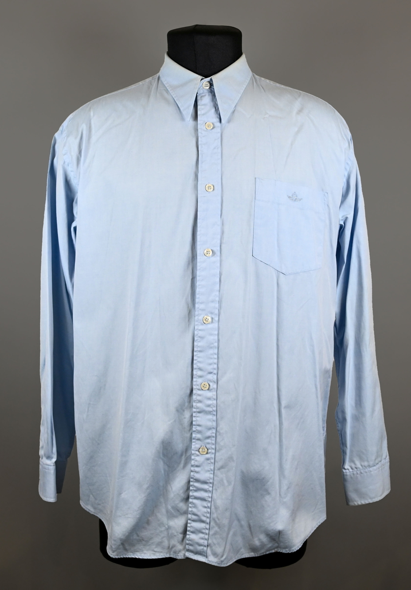 Blå långärmad skjorta som del av uniform från Luftfartsverket. Enkel knäppning med vita plastknappar och en bröstficka på vänster sida med Luftfartsverket logo broderad på. Vardera ärmslut har tre vita plastknappar vardera. I vänster innersöm titter en produktionsetikett med produktinformation och tvättråd. På etiketten är även två vita plastknappar i olika storlekar fastsydda.

Storlek L.