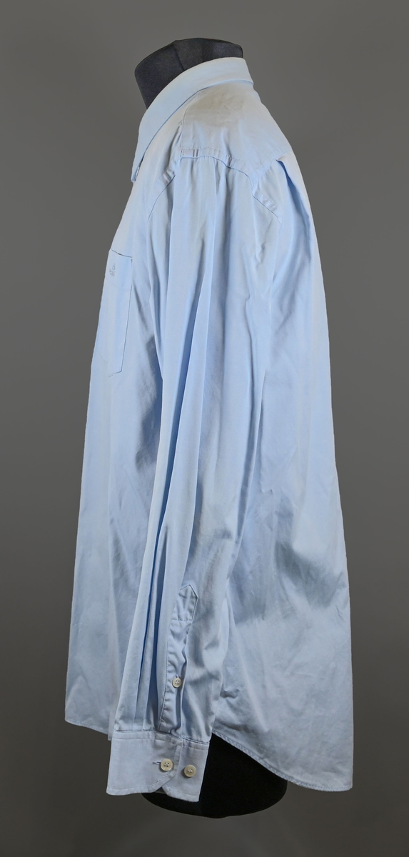 Blå långärmad skjorta som del av uniform från Luftfartsverket. Enkel knäppning med vita plastknappar och en bröstficka på vänster sida med Luftfartsverket logo broderad på. Vardera ärmslut har tre vita plastknappar vardera. I vänster innersöm titter en produktionsetikett med produktinformation och tvättråd. På etiketten är även två vita plastknappar i olika storlekar fastsydda.

Storlek L.