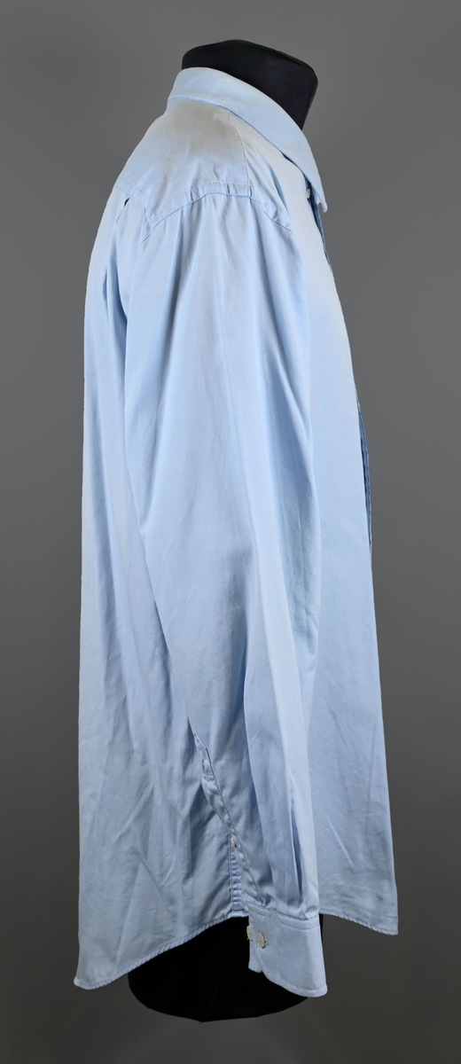 Blå långärmad skjorta som del av uniform från Luftfartsverket. Enkel knäppning med vita plastknappar och en bröstficka på vänster sida med Luftfartsverket logo broderad på. Vardera ärmslut har tre vita plastknappar vardera. I vänster innersöm titter en produktionsetikett med produktinformation och tvättråd. På etiketten är även två vita plastknappar i olika storlekar fastsydda.

Storlek L.