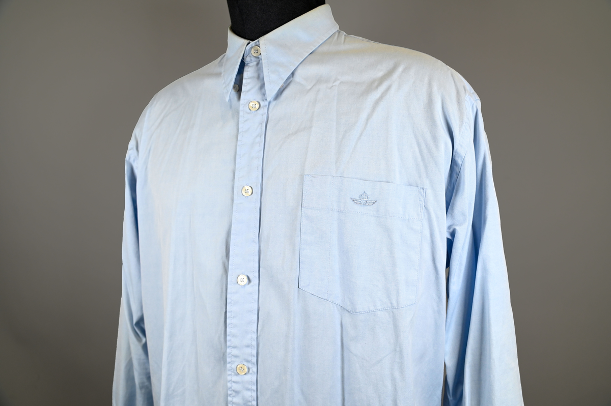 Blå långärmad skjorta som del av uniform från Luftfartsverket. Enkel knäppning med vita plastknappar och en bröstficka på vänster sida med Luftfartsverket logo broderad på. Vardera ärmslut har tre vita plastknappar vardera. I vänster innersöm titter en produktionsetikett med produktinformation och tvättråd. På etiketten är även två vita plastknappar i olika storlekar fastsydda.

Storlek L.