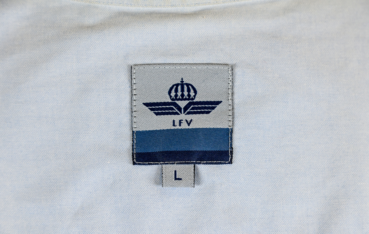 Blå långärmad skjorta som del av uniform från Luftfartsverket. Enkel knäppning med vita plastknappar och en bröstficka på vänster sida med Luftfartsverket logo broderad på. Vardera ärmslut har tre vita plastknappar vardera. I vänster innersöm titter en produktionsetikett med produktinformation och tvättråd. På etiketten är även två vita plastknappar i olika storlekar fastsydda.

Storlek L.