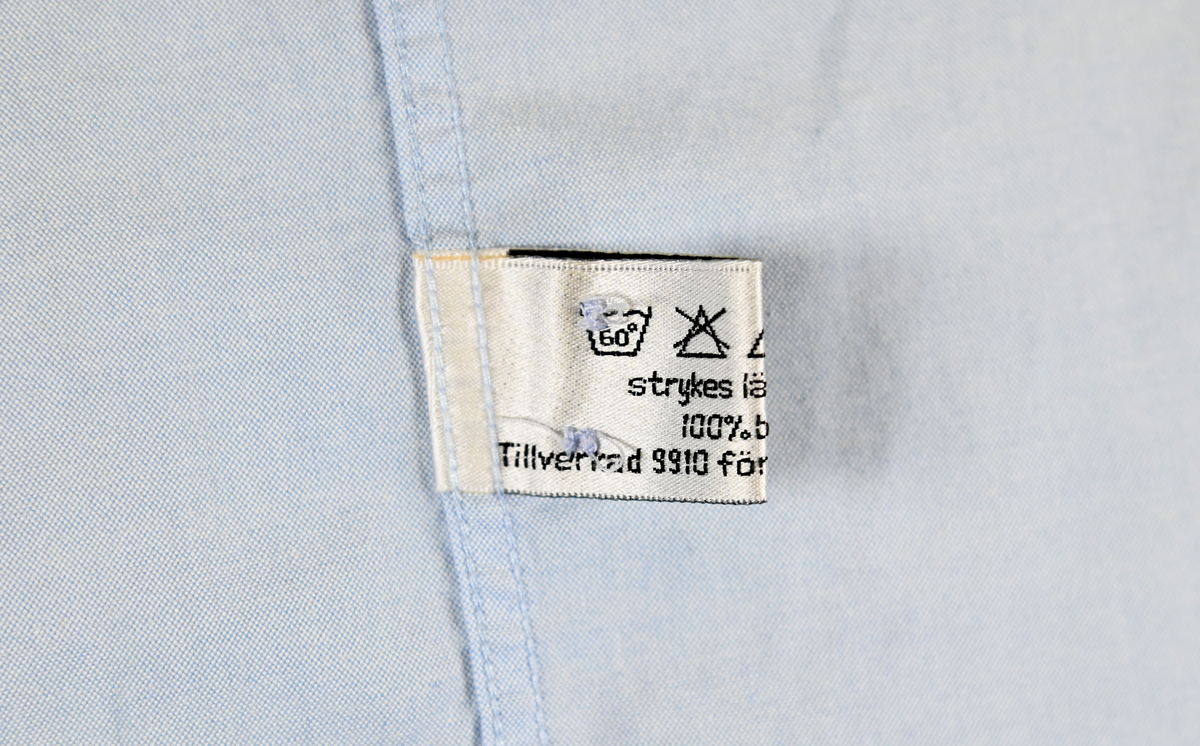 Blå långärmad skjorta som del av uniform från Luftfartsverket. Enkel knäppning med vita plastknappar och en bröstficka på vänster sida med Luftfartsverket logo broderad på. Vardera ärmslut har tre vita plastknappar vardera. I vänster innersöm titter en produktionsetikett med produktinformation och tvättråd. På etiketten är även två vita plastknappar i olika storlekar fastsydda.

Storlek L.