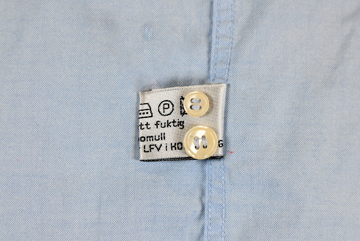 Blå långärmad skjorta som del av uniform från Luftfartsverket. Enkel knäppning med vita plastknappar och en bröstficka på vänster sida med Luftfartsverket logo broderad på. Vardera ärmslut har tre vita plastknappar vardera. I vänster innersöm titter en produktionsetikett med produktinformation och tvättråd. På etiketten är även två vita plastknappar i olika storlekar fastsydda.

Storlek L.