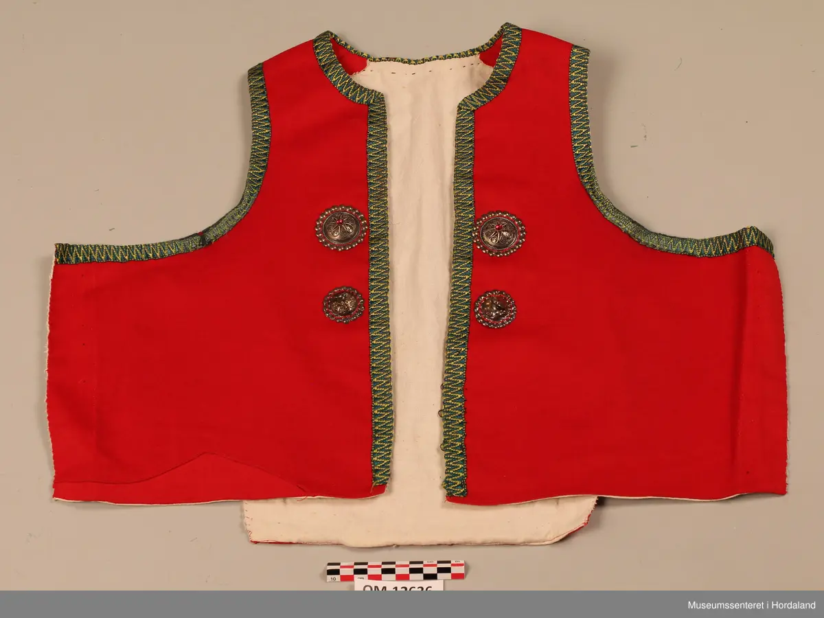 Raudt bunadsliv med grøne og gule silkekvardar (-band) rundt opningane og utanpå skuldrane (oppå skuldersaumen). Fire hekter i livet framme (kroker og maljer). To borer på kvar side langs opninga framme (ei stor og ei lita). Sidesaumane er spretta opp att. Ryggen er nokså smal så sidesaumane har vore trekte bakover. På eine framstykket er det skøytt på ein ekstra bit nede for å få rett fasong.