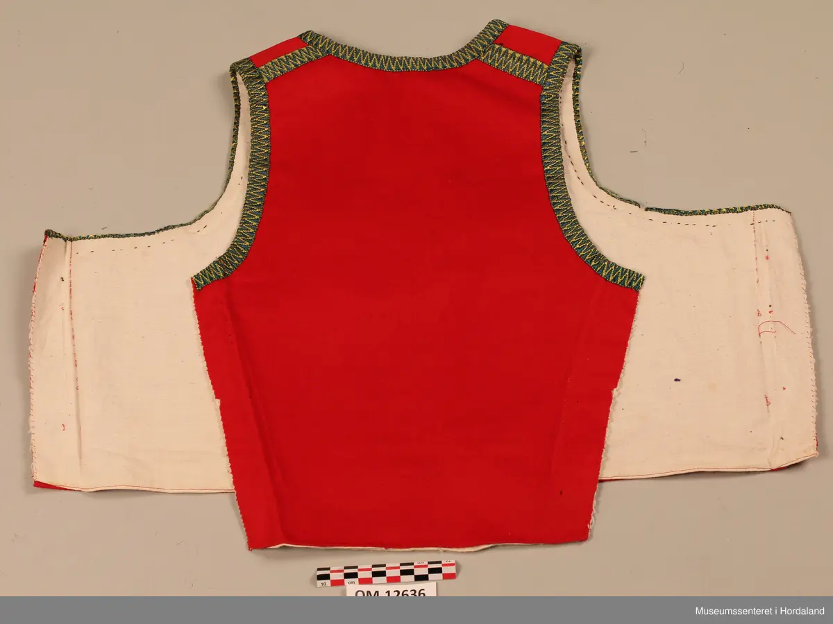 Raudt bunadsliv med grøne og gule silkekvardar (-band) rundt opningane og utanpå skuldrane (oppå skuldersaumen). Fire hekter i livet framme (kroker og maljer). To borer på kvar side langs opninga framme (ei stor og ei lita). Sidesaumane er spretta opp att. Ryggen er nokså smal så sidesaumane har vore trekte bakover. På eine framstykket er det skøytt på ein ekstra bit nede for å få rett fasong.