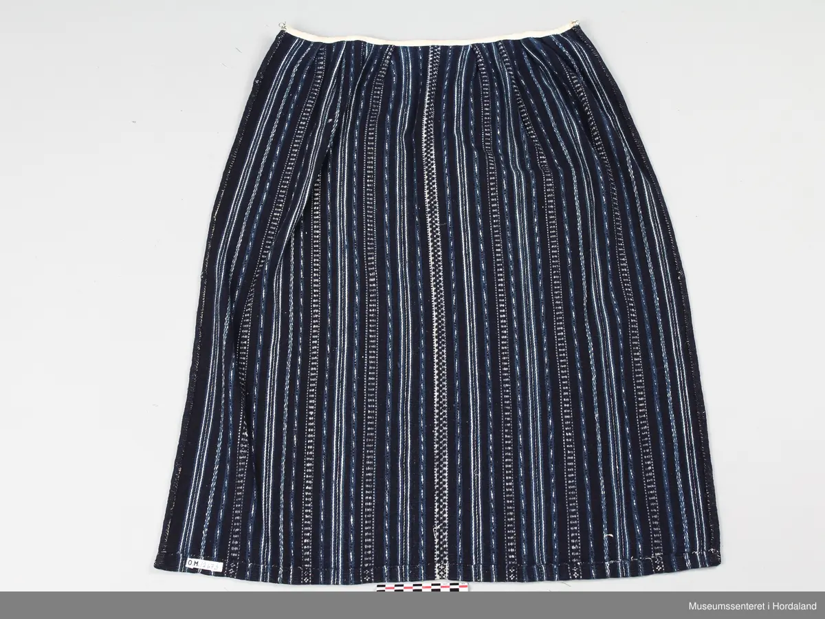 Vevd, mørkt forkle i tynt ullgarn med striper på langs. Stripene er i ulike teknikker så som krokbragd og rosebragd eller berre i enkle striper i innslagsrips slik som sjølve forkleet. Alle mønstra og stripene er nokså smale. Oppe i livet er det sydd på eit smalt kvitt bomullsband i staden for linning. Det har ei malje i kvar side. Innunder bomullsbandet er det 4 legg på kvar side.