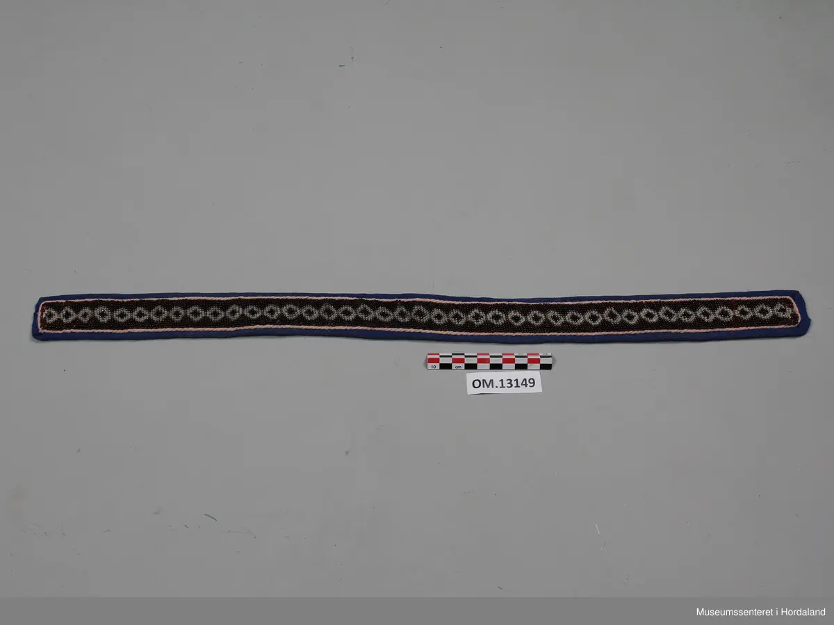 Perlebelte med broderte glassperler kanta med ullstoff. Perlearbeidet viser mønster med rundingar i gull, brunt og kvitt. Rundt perleborda er det ei tvinna, rosa snor på blå bakgrunn. Baksida av beltet er svart.