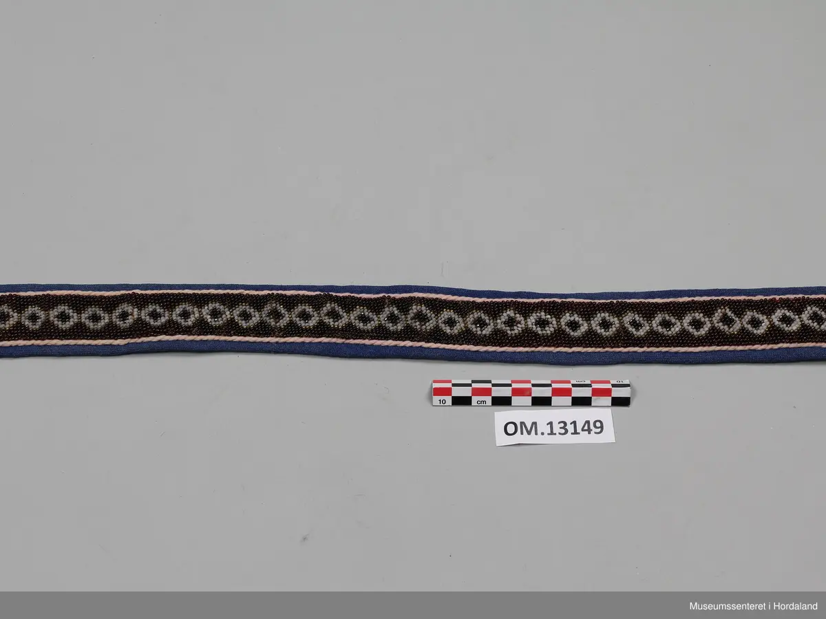 Perlebelte med broderte glassperler kanta med ullstoff. Perlearbeidet viser mønster med rundingar i gull, brunt og kvitt. Rundt perleborda er det ei tvinna, rosa snor på blå bakgrunn. Baksida av beltet er svart.