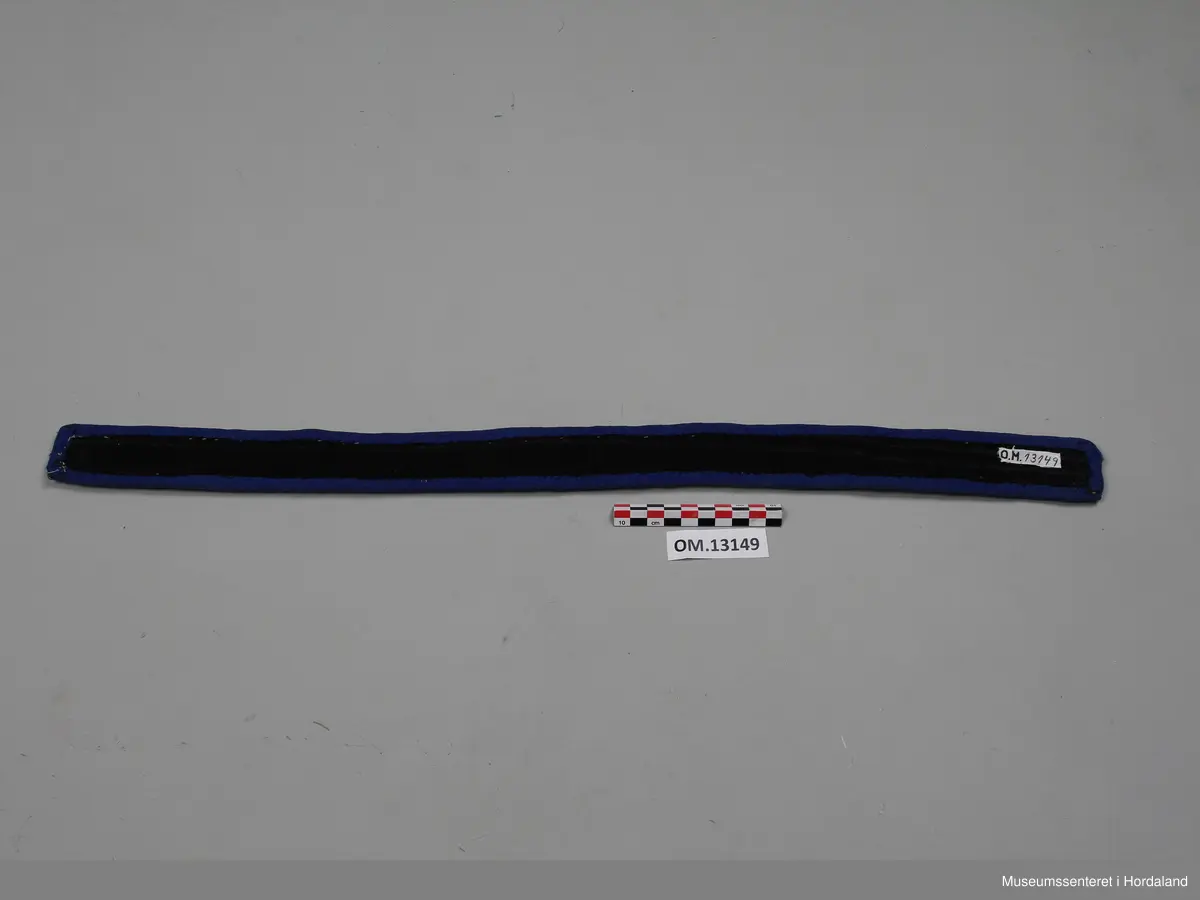 Perlebelte med broderte glassperler kanta med ullstoff. Perlearbeidet viser mønster med rundingar i gull, brunt og kvitt. Rundt perleborda er det ei tvinna, rosa snor på blå bakgrunn. Baksida av beltet er svart.