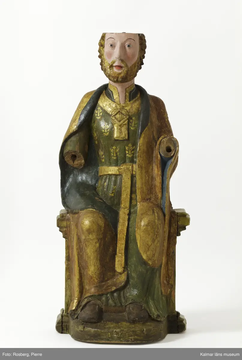 KLM 39475. Skulptur, kyrklig skulptur. Av lövträ. Föreställande en sittande Sankt Olof. Övre delen av hjässan är avsågad för att bereda underlag för ett offerfat eller kollektskål. På äldre bilder syns skulpturen ha ett platt kar/fat på hjässan. På huvudskulten finns också en inskärning för en krona. Bemålad i grön fotsid med förgyllda tryck. Skärp och halssmyckning är förgylld. Mantelns insida är målad i svart samt skorna, medans utsidan av manteln är förgylld. Saknar båda händer.