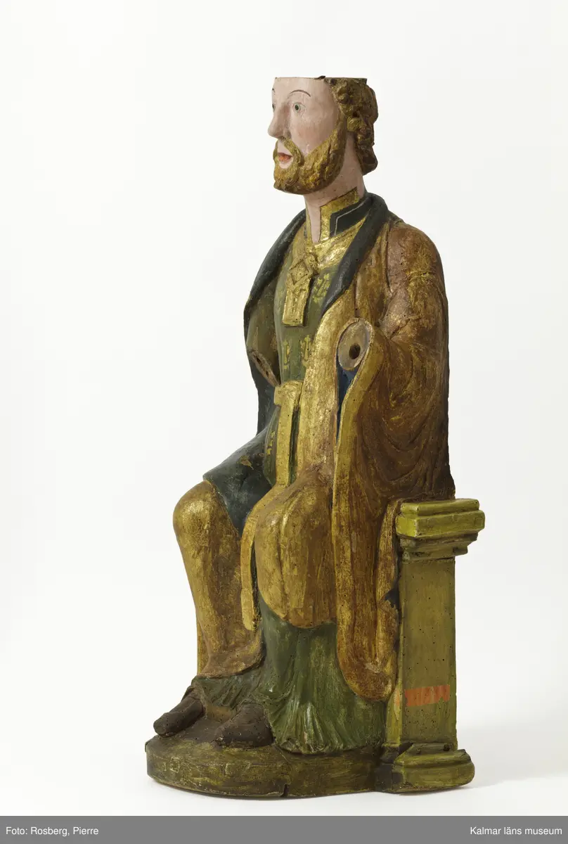 KLM 39475. Skulptur, kyrklig skulptur. Av lövträ. Föreställande en sittande Sankt Olof. Övre delen av hjässan är avsågad för att bereda underlag för ett offerfat eller kollektskål. På äldre bilder syns skulpturen ha ett platt kar/fat på hjässan. På huvudskulten finns också en inskärning för en krona. Bemålad i grön fotsid med förgyllda tryck. Skärp och halssmyckning är förgylld. Mantelns insida är målad i svart samt skorna, medans utsidan av manteln är förgylld. Saknar båda händer.