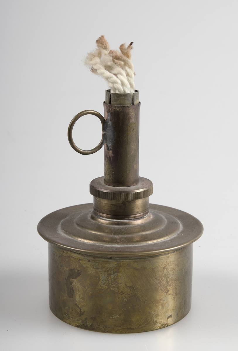Spritlampa av nysilver. Bomullsveke och ring som handtag. Cylinderformad behållare med rör upptill.