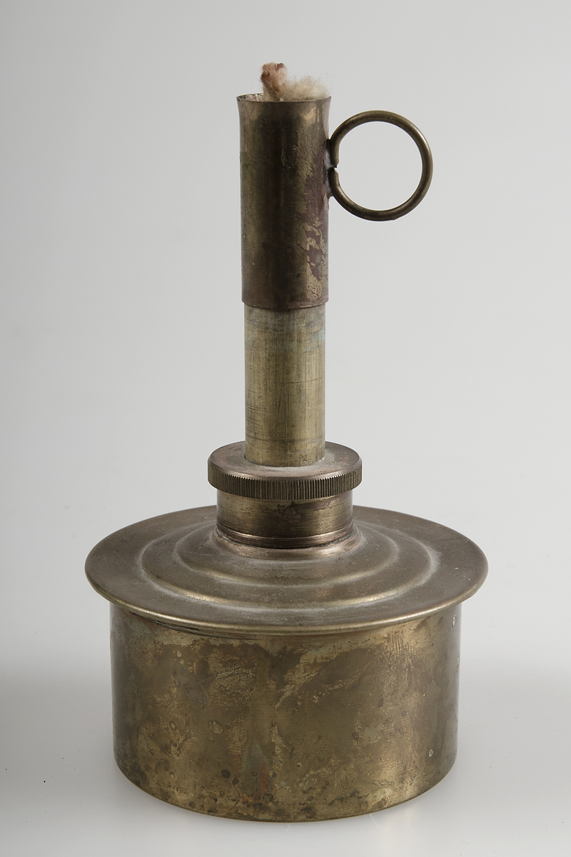 Spritlampa av nysilver. Bomullsveke och ring som handtag. Cylinderformad behållare med rör upptill.