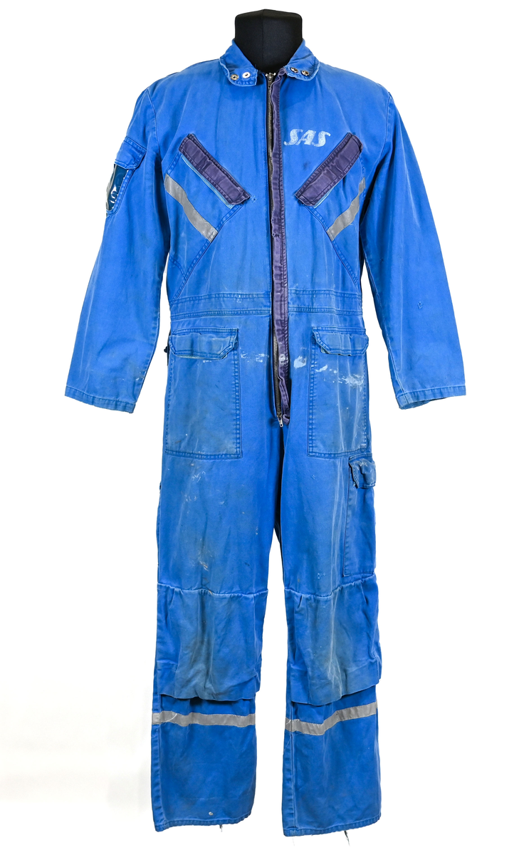 Blå arbetsoverall från SAS AB. På plagget finns en logga för SAS över vänster bröst och mitt på ryggen. Overallen har två bröstfickor med reflexband som försluts med kardborre. På framsidan under midjan sitter två större framfickor med lock och på vardera ben sitter ytterligare två fickor på sidorna. På baksidan finns två stora bakfickor. Högt upp på höger ärm sitter ytterligare en ficka av genomskinlig plast. Samtliga fickor försluts med kardborre förutom bakfickorna som stängs med plastknapp. Förutom på bröstfickorna sitter även reflexband runt benens nedre del samt högt upp över ryggen. Overallen sluts med dragkedja och två tryckknappar i metall på kragen. Vid höfterna finns två öppningar in i overallen. På knäna finns öppningar för att lägga i knäskydd.

Overallen är producerad av Fristads i storlek 50C. På lappen i nacken finns utrymme för namnteckning. På insidan av overallen vid midjan sitter en nött lapp som inte längre har någon information.

Plaggets framsida är befläckad med vit färg. Fotändar och ärmar är nötta. Flertalet mindre hål i höger ärm samt början till hål på vänster ärm.