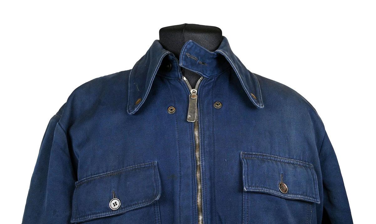 Mekanikeroverall i blå denim från Östgöta flygfottilj, F3. Overallen har två bröstfickor med lock som försluts med metallknapp. Längs ner vid fotändarna sitter en mindre ficka på vardera ben som sluts med dragkedja. På vardera sida vid midjan sitter fickor med dragkedja och löpare i skinn. På baksidan finns en bakficka på höger ben. Overallen har en krage som går att vika upp och stänga med två tygband och metallknappar. Den yttersta knappen på rad två av knappar sitter löst, Kragen går att vika ner och fästa i overallen med tryckknappar i metall. På insidan av midjan sitter en dragsko. Fotändarna kan spännas med en skinnrem. Overallen stängs med dragkedja i metall. 

På plaggets insida finns ett vitt tryck med texten "F3 2". F3 talar om att overallen användes vid Östgöta flygfottilj. 2:an anger troligtvis storleken på plagget.

Overallen har fyra stycken lagningar nere på höger baksida.