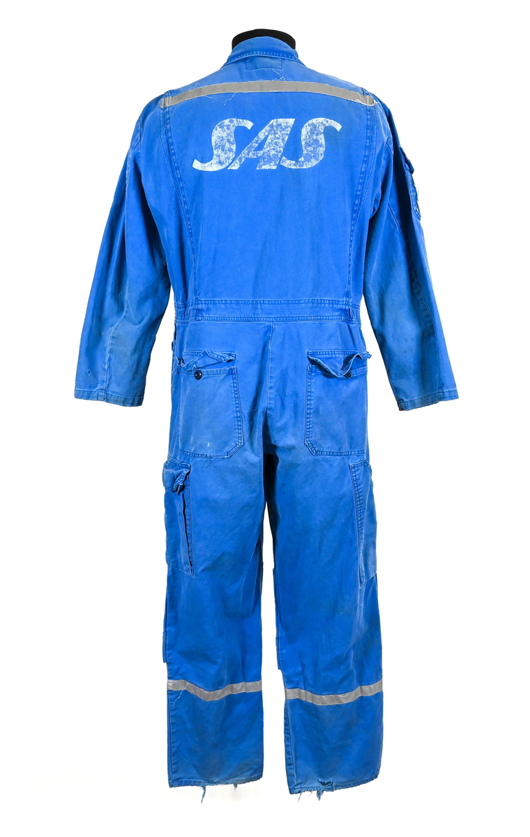 Blå arbetsoverall från SAS AB. På plagget finns en logga för SAS över vänster bröst och mitt på ryggen. Overallen har två bröstfickor med reflexband som försluts med kardborre. På framsidan under midjan sitter två större framfickor med lock och på vardera ben sitter ytterligare två fickor på sidorna. På baksidan finns två stora bakfickor. Högt upp på höger ärm sitter ytterligare en ficka av genomskinlig plast. Samtliga fickor försluts med kardborre förutom bakfickorna som stängs med plastknapp. Förutom på bröstfickorna sitter även reflexband runt benens nedre del samt högt upp över ryggen. Overallen sluts med dragkedja och två tryckknappar i metall på kragen. Vid höfterna finns två öppningar in i overallen. På knäna finns öppningar för att lägga i knäskydd.

Overallen är producerad av Fristads i storlek 50C. På lappen i nacken finns utrymme för namnteckning. På insidan av overallen vid midjan sitter en nött lapp som inte längre har någon information.

Plaggets framsida är befläckad med vit färg. Fotändar och ärmar är nötta. Flertalet mindre hål i höger ärm samt början till hål på vänster ärm.