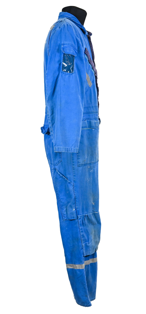 Blå arbetsoverall från SAS AB. På plagget finns en logga för SAS över vänster bröst och mitt på ryggen. Overallen har två bröstfickor med reflexband som försluts med kardborre. På framsidan under midjan sitter två större framfickor med lock och på vardera ben sitter ytterligare två fickor på sidorna. På baksidan finns två stora bakfickor. Högt upp på höger ärm sitter ytterligare en ficka av genomskinlig plast. Samtliga fickor försluts med kardborre förutom bakfickorna som stängs med plastknapp. Förutom på bröstfickorna sitter även reflexband runt benens nedre del samt högt upp över ryggen. Overallen sluts med dragkedja och två tryckknappar i metall på kragen. Vid höfterna finns två öppningar in i overallen. På knäna finns öppningar för att lägga i knäskydd.

Overallen är producerad av Fristads i storlek 50C. På lappen i nacken finns utrymme för namnteckning. På insidan av overallen vid midjan sitter en nött lapp som inte längre har någon information.

Plaggets framsida är befläckad med vit färg. Fotändar och ärmar är nötta. Flertalet mindre hål i höger ärm samt början till hål på vänster ärm.