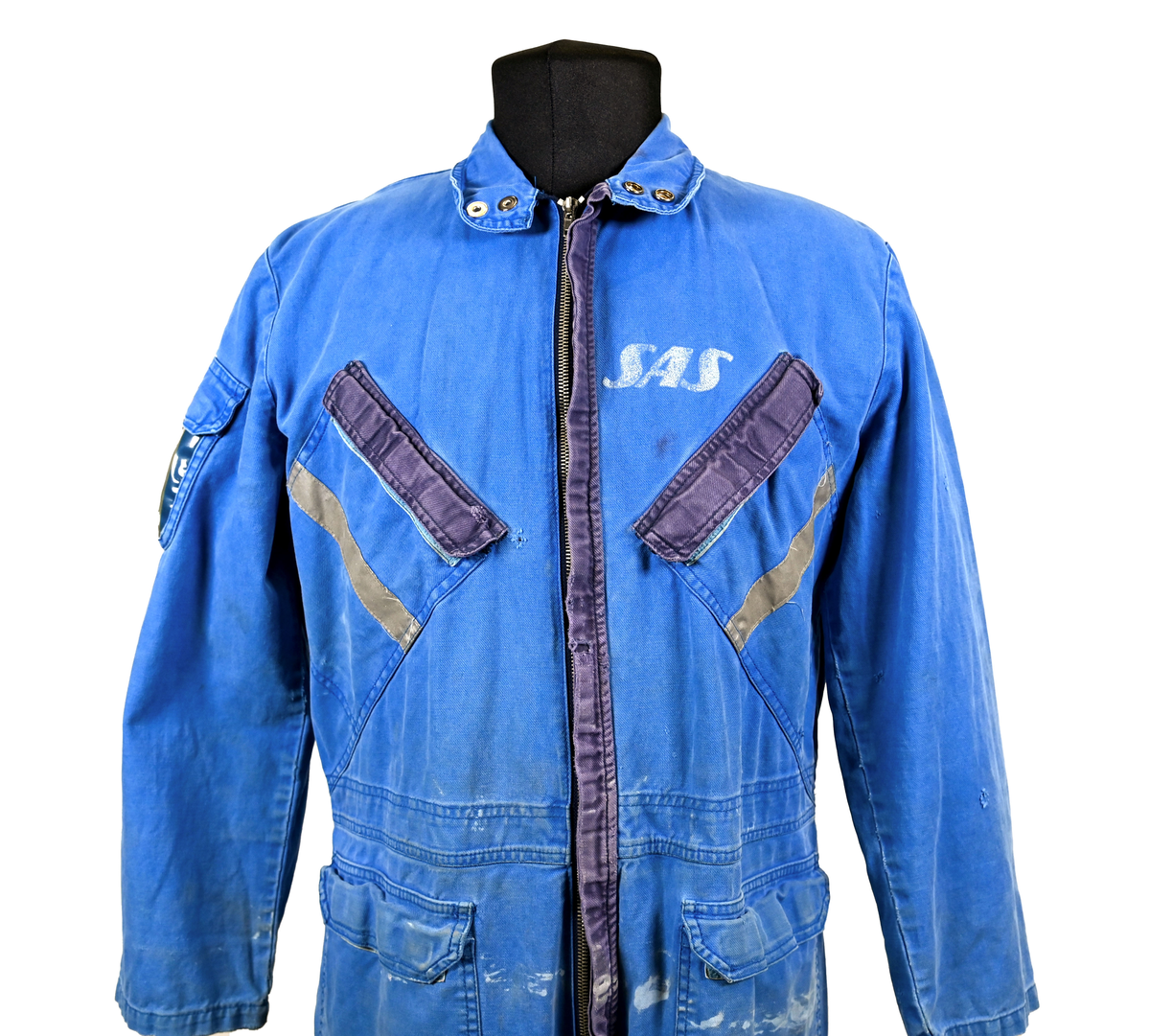 Blå arbetsoverall från SAS AB. På plagget finns en logga för SAS över vänster bröst och mitt på ryggen. Overallen har två bröstfickor med reflexband som försluts med kardborre. På framsidan under midjan sitter två större framfickor med lock och på vardera ben sitter ytterligare två fickor på sidorna. På baksidan finns två stora bakfickor. Högt upp på höger ärm sitter ytterligare en ficka av genomskinlig plast. Samtliga fickor försluts med kardborre förutom bakfickorna som stängs med plastknapp. Förutom på bröstfickorna sitter även reflexband runt benens nedre del samt högt upp över ryggen. Overallen sluts med dragkedja och två tryckknappar i metall på kragen. Vid höfterna finns två öppningar in i overallen. På knäna finns öppningar för att lägga i knäskydd.

Overallen är producerad av Fristads i storlek 50C. På lappen i nacken finns utrymme för namnteckning. På insidan av overallen vid midjan sitter en nött lapp som inte längre har någon information.

Plaggets framsida är befläckad med vit färg. Fotändar och ärmar är nötta. Flertalet mindre hål i höger ärm samt början till hål på vänster ärm.