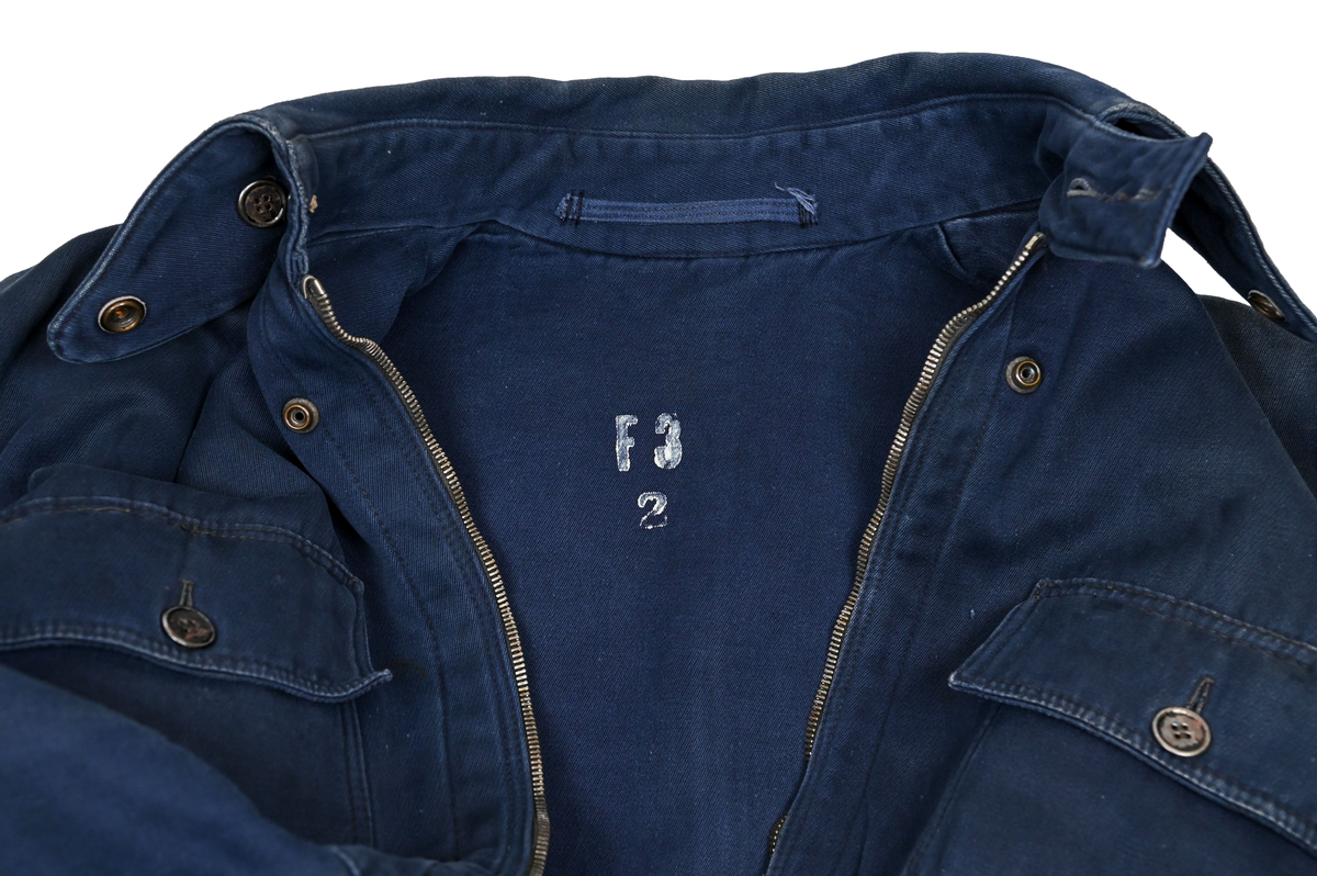 Mekanikeroverall i blå denim från Östgöta flygfottilj, F3. Overallen har två bröstfickor med lock som försluts med metallknapp. Längs ner vid fotändarna sitter en mindre ficka på vardera ben som sluts med dragkedja. På vardera sida vid midjan sitter fickor med dragkedja och löpare i skinn. På baksidan finns en bakficka på höger ben. Overallen har en krage som går att vika upp och stänga med två tygband och metallknappar. Den yttersta knappen på rad två av knappar sitter löst, Kragen går att vika ner och fästa i overallen med tryckknappar i metall. På insidan av midjan sitter en dragsko. Fotändarna kan spännas med en skinnrem. Overallen stängs med dragkedja i metall. 

På plaggets insida finns ett vitt tryck med texten "F3 2". F3 talar om att overallen användes vid Östgöta flygfottilj. 2:an anger troligtvis storleken på plagget.

Overallen har fyra stycken lagningar nere på höger baksida.