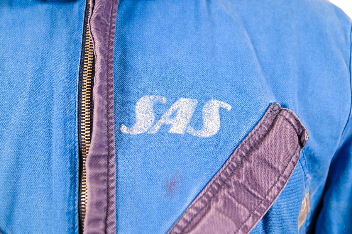 Blå arbetsoverall från SAS AB. På plagget finns en logga för SAS över vänster bröst och mitt på ryggen. Overallen har två bröstfickor med reflexband som försluts med kardborre. På framsidan under midjan sitter två större framfickor med lock och på vardera ben sitter ytterligare två fickor på sidorna. På baksidan finns två stora bakfickor. Högt upp på höger ärm sitter ytterligare en ficka av genomskinlig plast. Samtliga fickor försluts med kardborre förutom bakfickorna som stängs med plastknapp. Förutom på bröstfickorna sitter även reflexband runt benens nedre del samt högt upp över ryggen. Overallen sluts med dragkedja och två tryckknappar i metall på kragen. Vid höfterna finns två öppningar in i overallen. På knäna finns öppningar för att lägga i knäskydd.

Overallen är producerad av Fristads i storlek 50C. På lappen i nacken finns utrymme för namnteckning. På insidan av overallen vid midjan sitter en nött lapp som inte längre har någon information.

Plaggets framsida är befläckad med vit färg. Fotändar och ärmar är nötta. Flertalet mindre hål i höger ärm samt början till hål på vänster ärm.