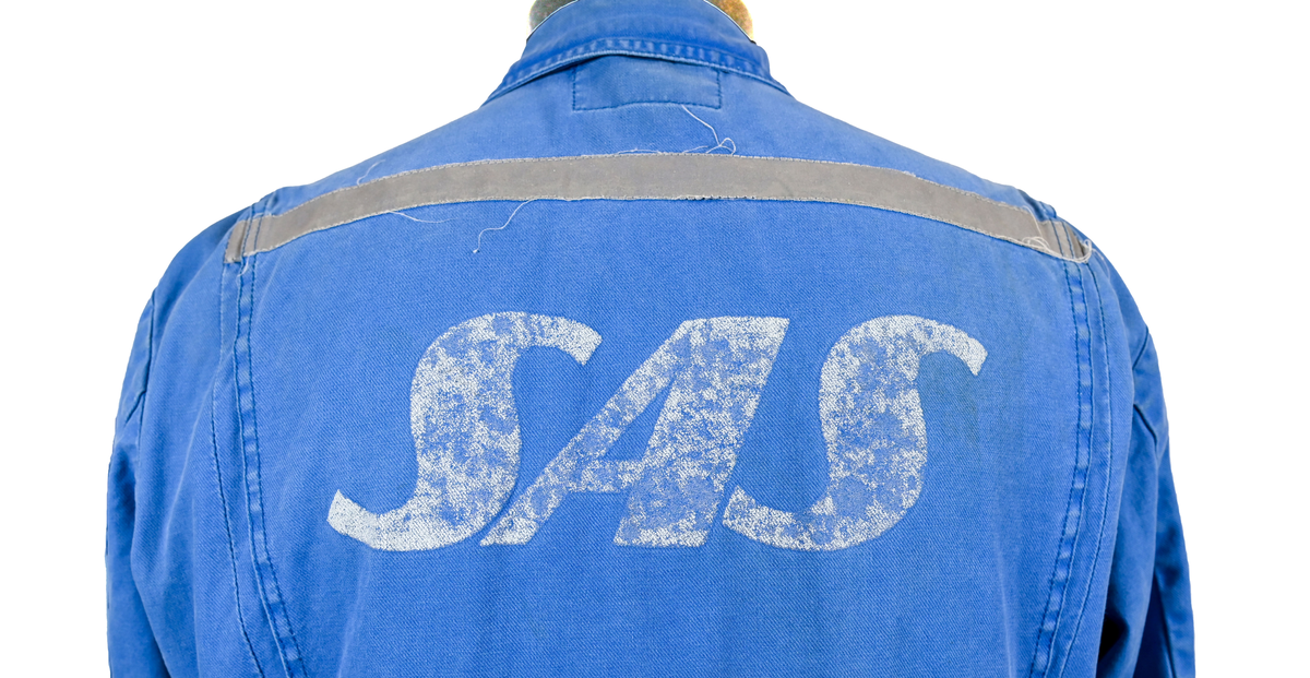 Blå arbetsoverall från SAS AB. På plagget finns en logga för SAS över vänster bröst och mitt på ryggen. Overallen har två bröstfickor med reflexband som försluts med kardborre. På framsidan under midjan sitter två större framfickor med lock och på vardera ben sitter ytterligare två fickor på sidorna. På baksidan finns två stora bakfickor. Högt upp på höger ärm sitter ytterligare en ficka av genomskinlig plast. Samtliga fickor försluts med kardborre förutom bakfickorna som stängs med plastknapp. Förutom på bröstfickorna sitter även reflexband runt benens nedre del samt högt upp över ryggen. Overallen sluts med dragkedja och två tryckknappar i metall på kragen. Vid höfterna finns två öppningar in i overallen. På knäna finns öppningar för att lägga i knäskydd.

Overallen är producerad av Fristads i storlek 50C. På lappen i nacken finns utrymme för namnteckning. På insidan av overallen vid midjan sitter en nött lapp som inte längre har någon information.

Plaggets framsida är befläckad med vit färg. Fotändar och ärmar är nötta. Flertalet mindre hål i höger ärm samt början till hål på vänster ärm.