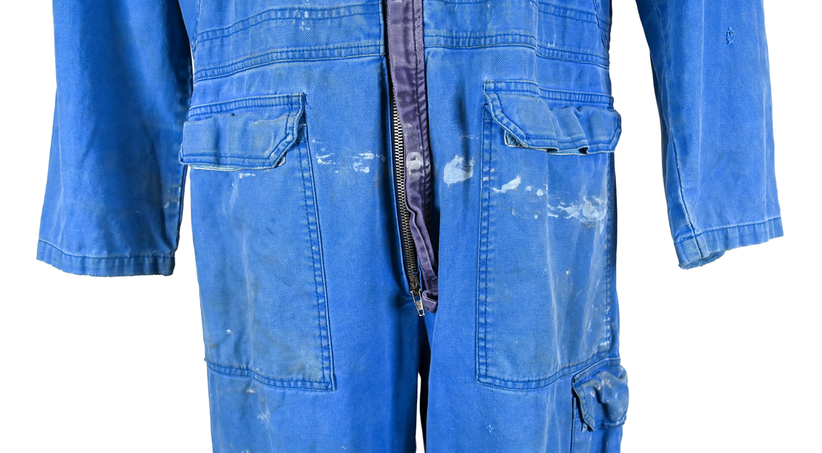 Blå arbetsoverall från SAS AB. På plagget finns en logga för SAS över vänster bröst och mitt på ryggen. Overallen har två bröstfickor med reflexband som försluts med kardborre. På framsidan under midjan sitter två större framfickor med lock och på vardera ben sitter ytterligare två fickor på sidorna. På baksidan finns två stora bakfickor. Högt upp på höger ärm sitter ytterligare en ficka av genomskinlig plast. Samtliga fickor försluts med kardborre förutom bakfickorna som stängs med plastknapp. Förutom på bröstfickorna sitter även reflexband runt benens nedre del samt högt upp över ryggen. Overallen sluts med dragkedja och två tryckknappar i metall på kragen. Vid höfterna finns två öppningar in i overallen. På knäna finns öppningar för att lägga i knäskydd.

Overallen är producerad av Fristads i storlek 50C. På lappen i nacken finns utrymme för namnteckning. På insidan av overallen vid midjan sitter en nött lapp som inte längre har någon information.

Plaggets framsida är befläckad med vit färg. Fotändar och ärmar är nötta. Flertalet mindre hål i höger ärm samt början till hål på vänster ärm.