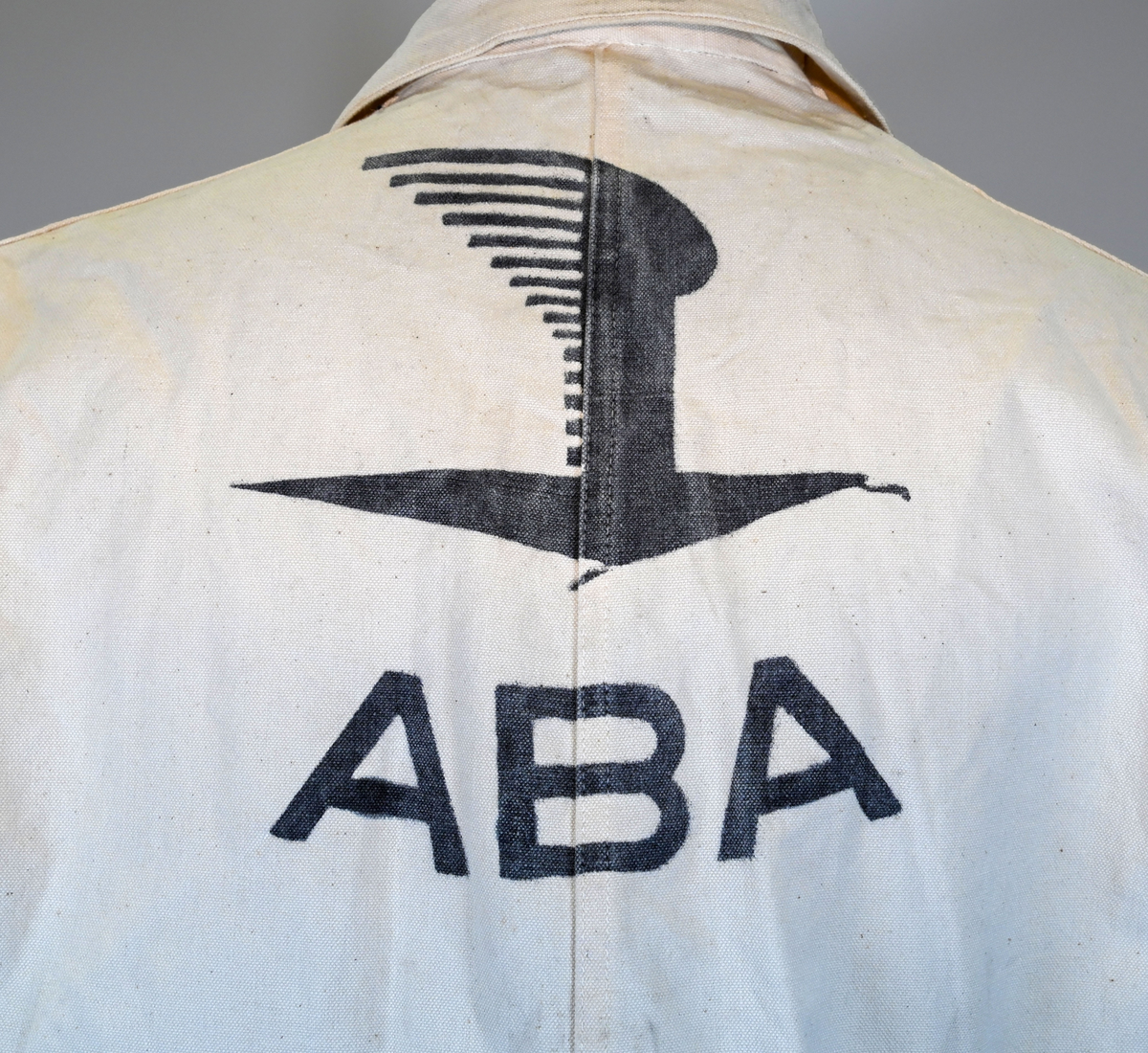 Arbetsoverall i vitt, grövre tyg för personal vid AB Aerotransport. På ryggen finns ett tryck i mörkgrå färg föreställande företagets logga och initialer, ABA. Overallen har två bröstfickor på vardera sida samt två fram- och bakfickor. Under bakfickan på höger ben sitter ytterligare en mindre ficka. Overallen har krage och mässingsfärgade metallknappar. Plagget är utsatt för visst slitage och smuts.