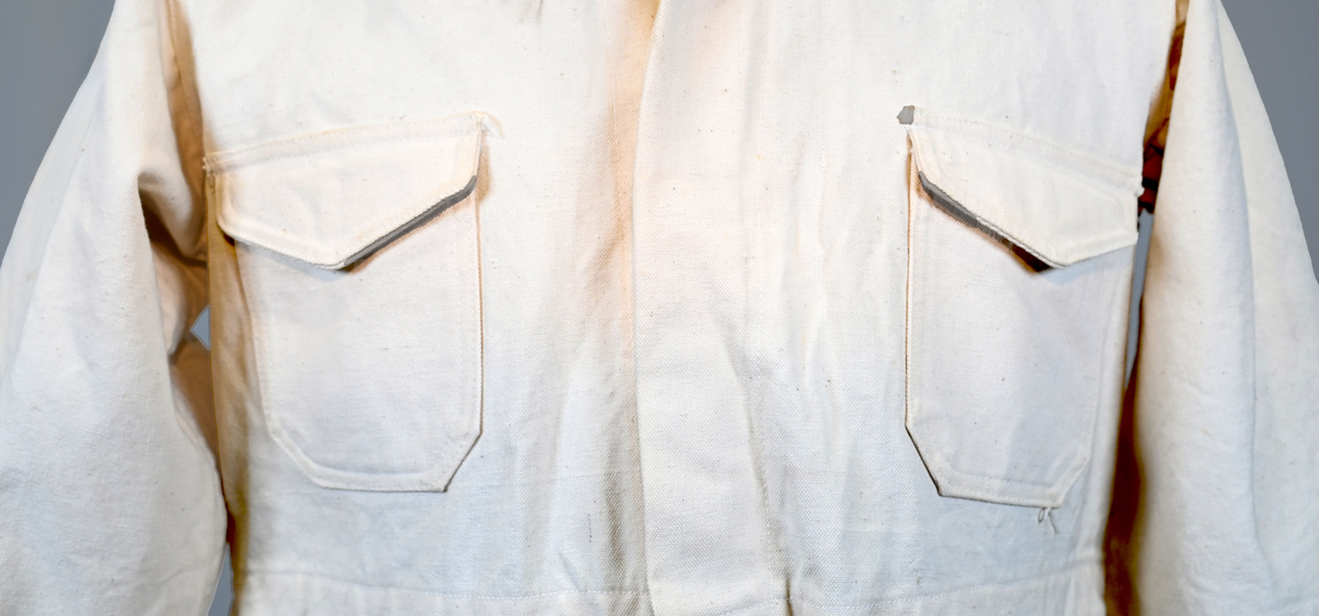 Arbetsoverall i vitt, grövre tyg för personal vid AB Aerotransport. På ryggen finns ett tryck i mörkgrå färg föreställande företagets logga och initialer, ABA. Overallen har två bröstfickor på vardera sida samt två fram- och bakfickor. Under bakfickan på höger ben sitter ytterligare en mindre ficka. Overallen har krage och mässingsfärgade metallknappar. Plagget är utsatt för visst slitage och smuts.