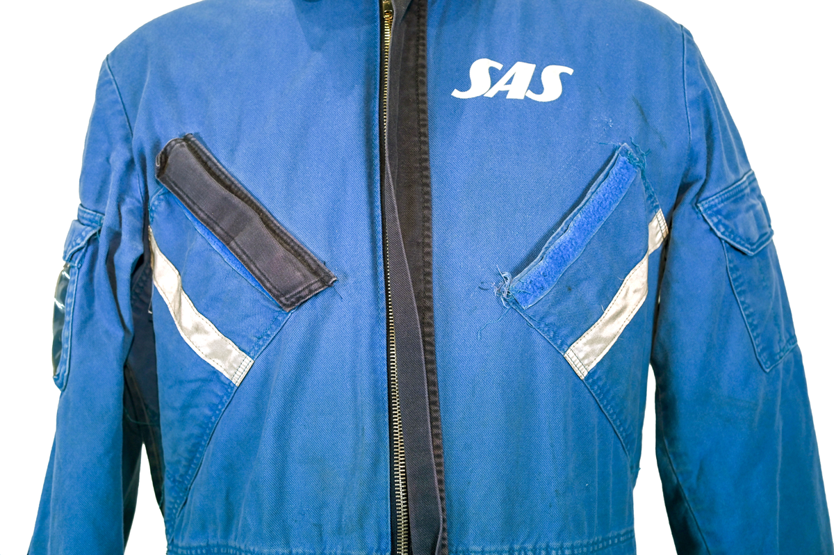 Blå arbetsoverall från SAS. På plagget finns en logga för SAS över vänster bröst och mitt på ryggen. Overallen har två bröstfickor med reflexband som försluts med kardborre. Vänster ficka saknar den del som kopplar an till kardborren på fickan. Frampå sitter två större framfickor med lock och på vardera ben sitter ytterligare två fickor på sidorna. På baksidan finns två stora bakfickor. Högt upp på sidan av vardera ärm sitter ytterligare fickor varav den högra är av genomskinlig plast. Samtliga fickor försluts med kardborre. Utöver på bröstfickorna sitter reflexband runt benens nedre del samt högt upp över ryggen. Overallen sluts med dragkedja och två tryckknappar i metall. Ärmsluten kan justeras med kardborre. Vid höfterna finns två öppningar in i overallen. På knäna finns öppningar för att lägga i knäskydd.

På plagget finns spår av vit färg och andra smutsfläckar. Vänster bakficka är nött.

Overallen är producerad av Fristads i storlek 48. På insidan längs med dragskedjan sitter en lapp med texten "OBS! DRAGKEDJAN SKALL VARA STÄNGD VID TVÄTT".