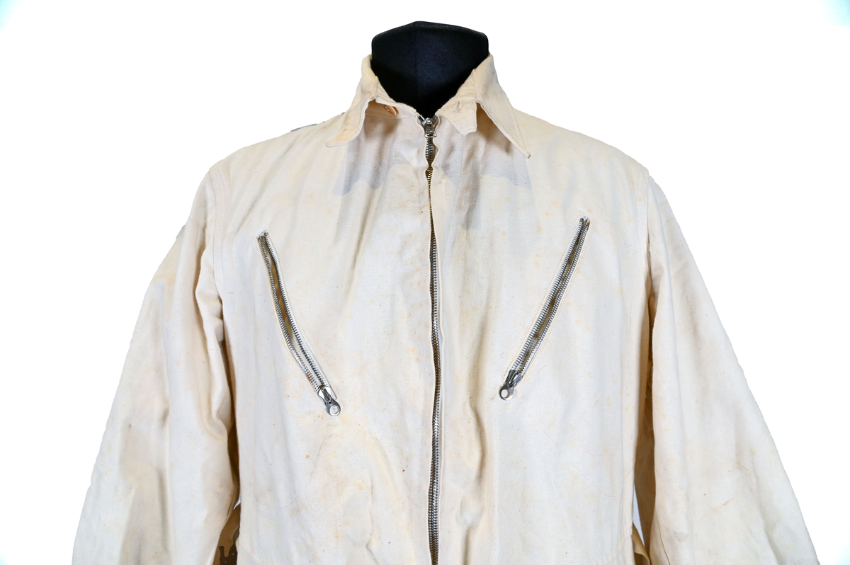 Långärmad overall i vitt tyg med krage. Overallen har två bröstfickor och två framfickor som sluts med dragkedja i metall. I midjan sitter ett bälte i samma material som plagget med ett spänne i metall. Bältet sitter på plats med med två öglor i plaggets sidor. Ärmsluten kan knäppas med tygband och knappar i plast. Bensluten kan spännas med tygband fäst i fyra öglor med metallspänne. 

Overallen är täckt av smuts och fläckar.