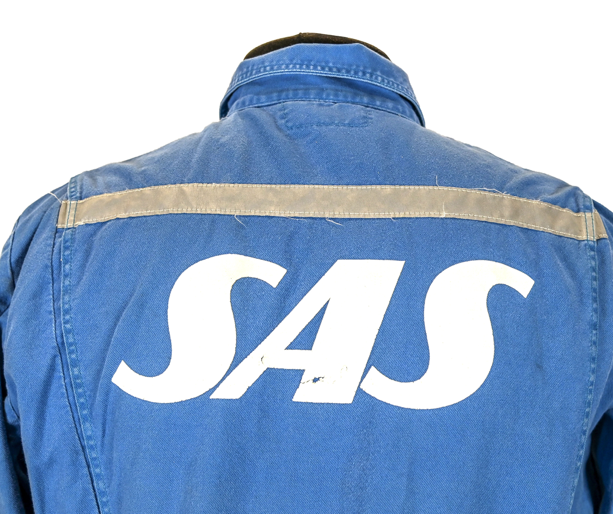 Blå arbetsoverall från SAS. På plagget finns en logga för SAS över vänster bröst och mitt på ryggen. Overallen har två bröstfickor med reflexband som försluts med kardborre. Vänster ficka saknar den del som kopplar an till kardborren på fickan. Frampå sitter två större framfickor med lock och på vardera ben sitter ytterligare två fickor på sidorna. På baksidan finns två stora bakfickor. Högt upp på sidan av vardera ärm sitter ytterligare fickor varav den högra är av genomskinlig plast. Samtliga fickor försluts med kardborre. Utöver på bröstfickorna sitter reflexband runt benens nedre del samt högt upp över ryggen. Overallen sluts med dragkedja och två tryckknappar i metall. Ärmsluten kan justeras med kardborre. Vid höfterna finns två öppningar in i overallen. På knäna finns öppningar för att lägga i knäskydd.

På plagget finns spår av vit färg och andra smutsfläckar. Vänster bakficka är nött.

Overallen är producerad av Fristads i storlek 48. På insidan längs med dragskedjan sitter en lapp med texten "OBS! DRAGKEDJAN SKALL VARA STÄNGD VID TVÄTT".