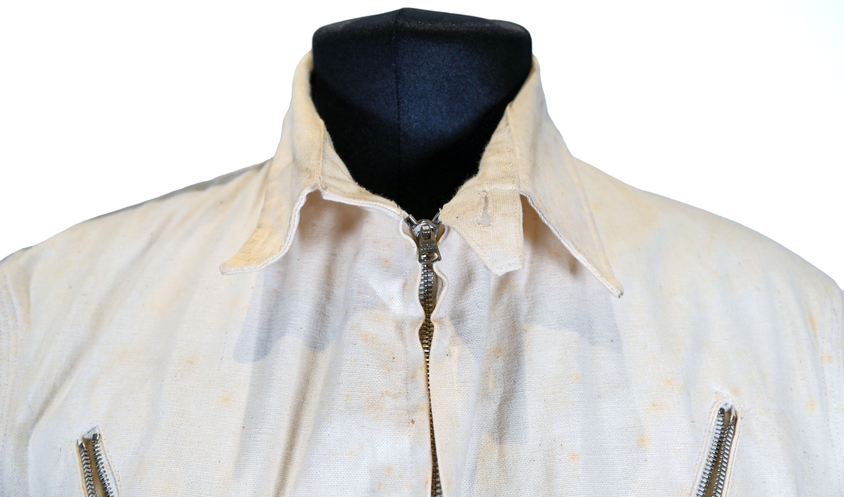 Långärmad overall i vitt tyg med krage. Overallen har två bröstfickor och två framfickor som sluts med dragkedja i metall. I midjan sitter ett bälte i samma material som plagget med ett spänne i metall. Bältet sitter på plats med med två öglor i plaggets sidor. Ärmsluten kan knäppas med tygband och knappar i plast. Bensluten kan spännas med tygband fäst i fyra öglor med metallspänne. 

Overallen är täckt av smuts och fläckar.