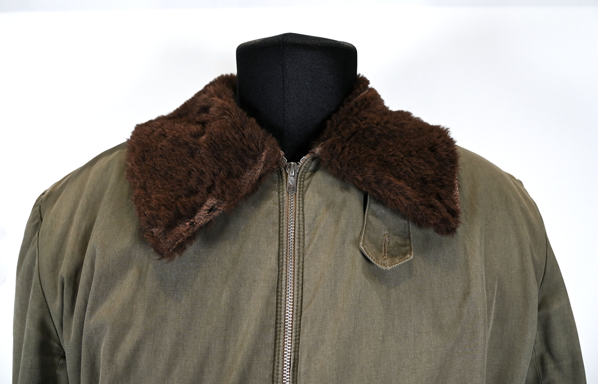 Grågrön overall som använts som flygardräkt. Liknande dräkter har använts inom flygvapnet. Dräkten är fodrad och har en fodrad krage i brun päls. I ärmsluten sitter en mörkblå resår. På framsida lår sitter en ficka på vardera ben som försluts med dragkedja i metall. På sidorna i midjan sitter en öppning som sluts med dragkedja. Dräkten stängs med en lång dragkedja i metall som sträcker sig ned i höger ben. I kragen sitter ett tygband som stänger igen kragen med en plastknapp.  Vid fotändarna finns dragkedja och knapp för att öppna upp fotsluten. 

I tygbandet på kragen satt (borttagen och lagd i ficka av vårdskäl) en lapp fasthäftad som troligtvis har fästs i plagget i samband med tvätt.