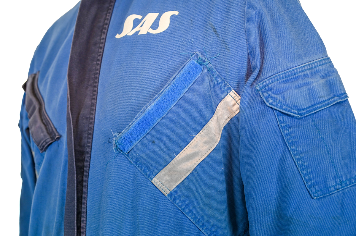 Blå arbetsoverall från SAS. På plagget finns en logga för SAS över vänster bröst och mitt på ryggen. Overallen har två bröstfickor med reflexband som försluts med kardborre. Vänster ficka saknar den del som kopplar an till kardborren på fickan. Frampå sitter två större framfickor med lock och på vardera ben sitter ytterligare två fickor på sidorna. På baksidan finns två stora bakfickor. Högt upp på sidan av vardera ärm sitter ytterligare fickor varav den högra är av genomskinlig plast. Samtliga fickor försluts med kardborre. Utöver på bröstfickorna sitter reflexband runt benens nedre del samt högt upp över ryggen. Overallen sluts med dragkedja och två tryckknappar i metall. Ärmsluten kan justeras med kardborre. Vid höfterna finns två öppningar in i overallen. På knäna finns öppningar för att lägga i knäskydd.

På plagget finns spår av vit färg och andra smutsfläckar. Vänster bakficka är nött.

Overallen är producerad av Fristads i storlek 48. På insidan längs med dragskedjan sitter en lapp med texten "OBS! DRAGKEDJAN SKALL VARA STÄNGD VID TVÄTT".